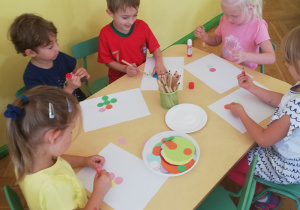 Zdjęcie przedstawia pięcioro dzieci siedzących przy stoliku przyklejających kolorowe kropki na białym kartonie
